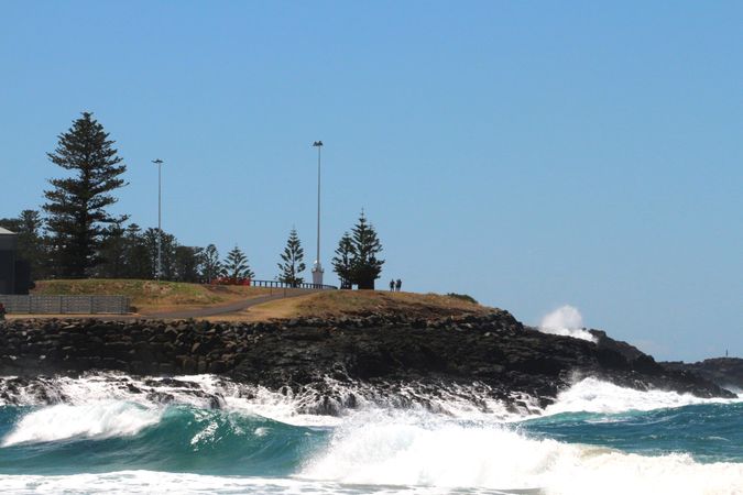 Waves crash against rocks near a coastal path with visitors enjoying the sunny day at the shoreline