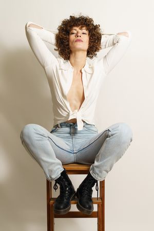 Woman in jeans and boots sitting on wooden stool in studio shot looking at camera