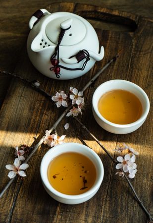 Cups of green tea and pot with floral petals on wooden board