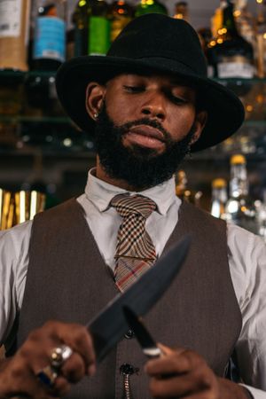 Close-up of a stylish Black bartender in a hat sharpening his knives