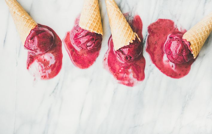Four cones of dark berry ice cream melting on marble slab, horizontal composition with copy space