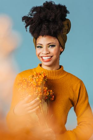 Studio shoot of Black woman holding orange flowers behind orange cloud effect in a blue room