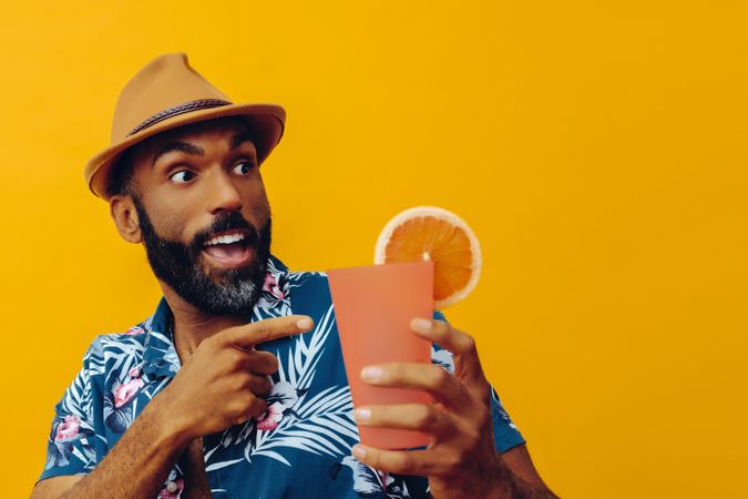 Surprised male wearing colorful shirt and pointing at cocktail with yellow background