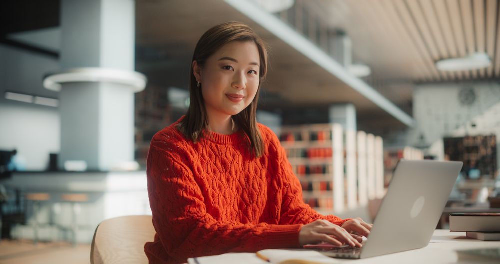 Confident Asian Female Student Working on a Laptop in a Modern Library.