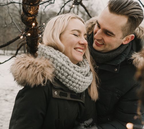 Smiling couple having fun on street near tree with fairy lights
