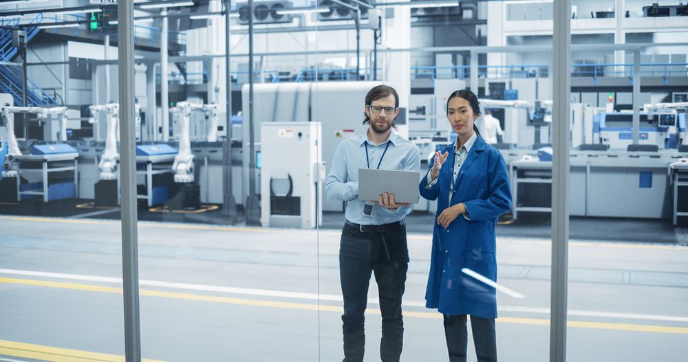 Man and Asian Female Having a Conversation at an Industrial Facility. Advanced Factory Employees Working Together, Using Laptop Computer for Supervising Work Process in a Modern Facility