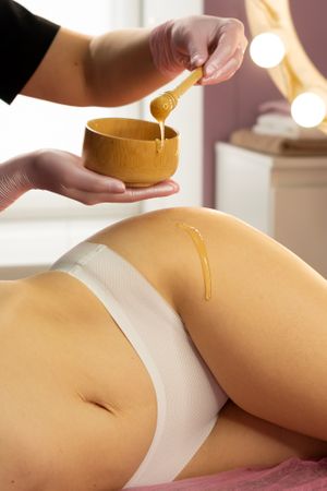 Beautician dripping honey onto thigh during cosmetic body treatment.
