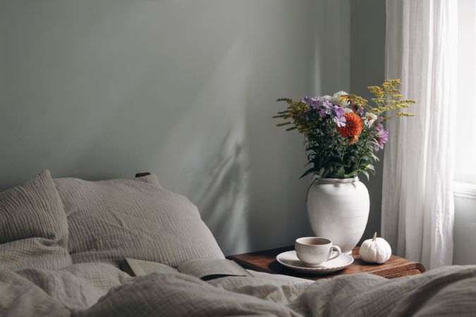 Cup of coffee, pumpkin on retro wooden bedside table, bouquet of dahlia, cosmos, solidago flowers, beige muslin cushions, book in bed near window