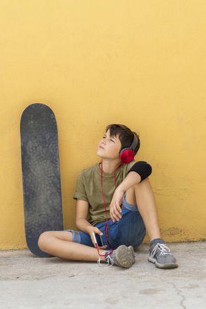 Teenager sitting on ground leaning on a yellow wall while holding a mobile phone looking up