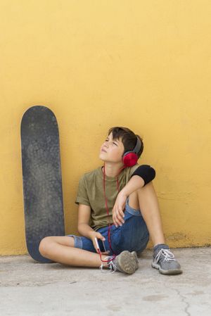 Teenager sitting on ground leaning on a yellow wall while holding a mobile phone looking up