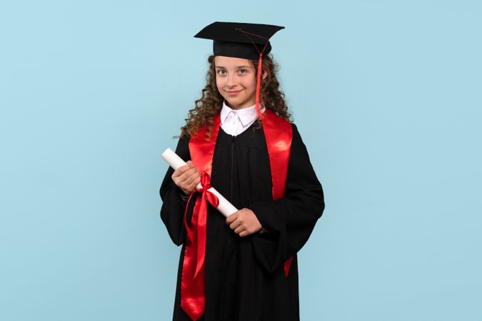 Young girl in graduation attire holding diploma and smiling confidently.