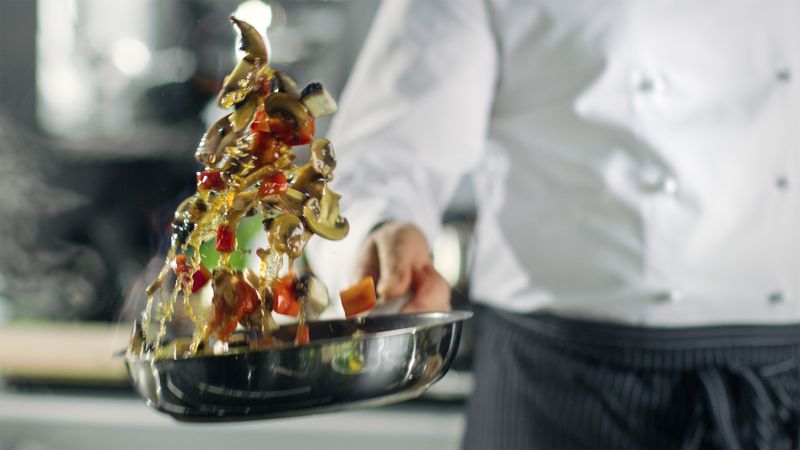 Professional Chef Cooks Flambe Style. He Prepares Dish in a Pan with Open Flames. He Works in a Modern Kitchen with Different Ingredients Lying Around.