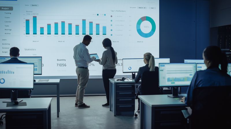 Data Analysts Collaborate in Dimly Lit Office, Examining Project Metrics on Large Screen.