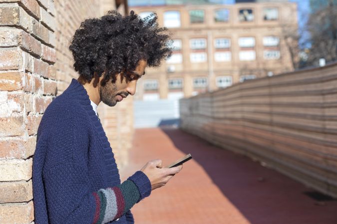 Curly haired male looking down at his smartphone while leaning on a brick wall outdoors on sunny day