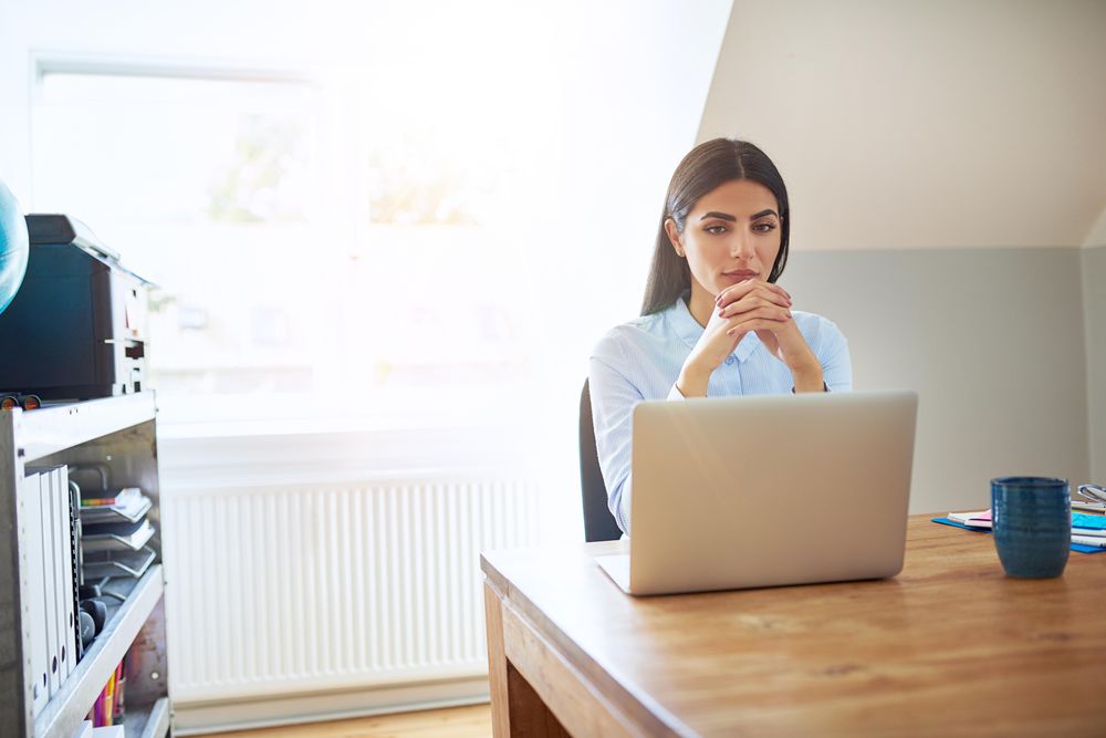 Pensive woman sitting at her laptop reading screen with hands clasped deep in thought