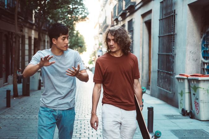 Cool multiracial male friends chatting while walking on street with longboard