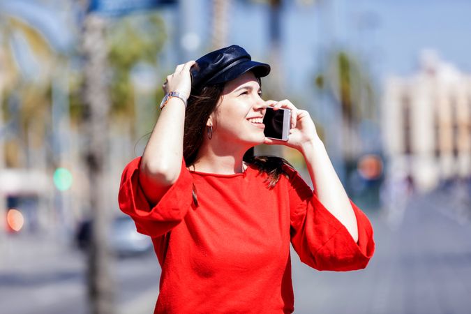 Young woman wearing red blouse sitting on a metallic fence while using a mobile phone outdoors in the street on a bright day