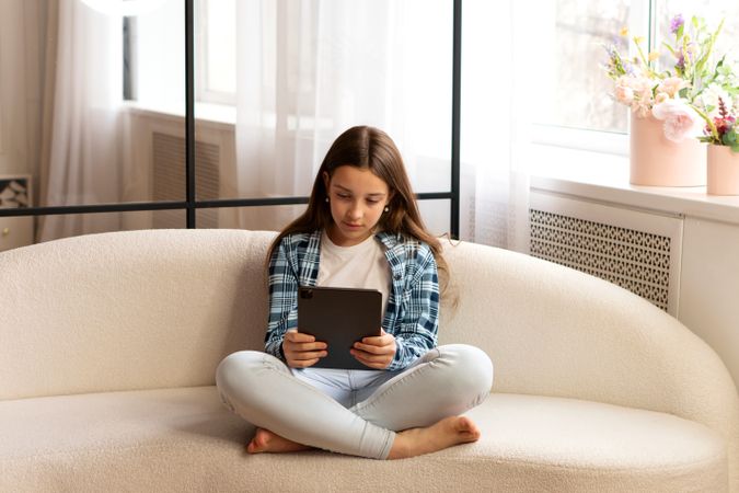 Girl sitting cross-legged on sofa using tablet at home.