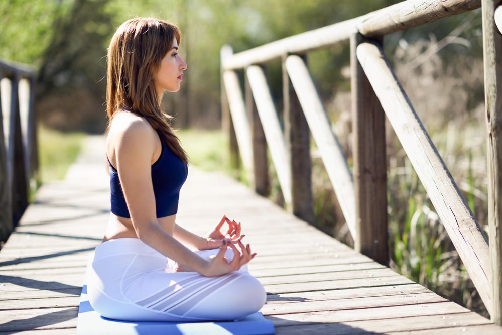 Side view of woman wearing sport clothes meditating with eyes closed on yoga mat on forest path