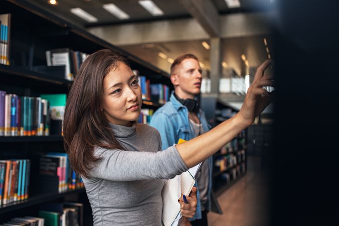 Young female student selecting book from library shelf with man in background