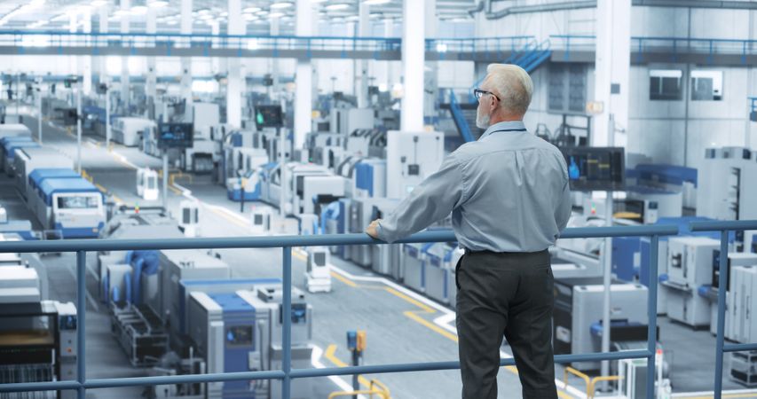 Industrial Engineer Working at a Technology Factory. Mature Manager Standing on a Platform at Work, Looking at the Production Manufacturing Complex. Automated Robots Moving Around the Hall
