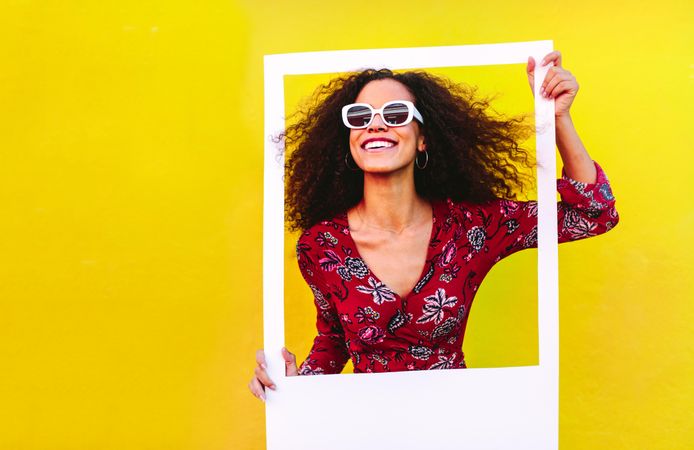 Woman with curly hair wearing sundress and sunglasses looking through blank photo frame