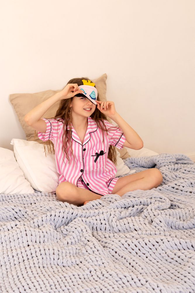 Girl playfully adjusting sleep mask while sitting on bed.