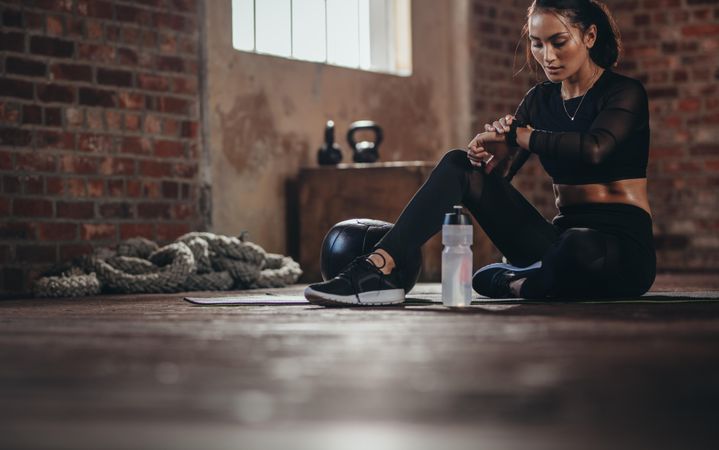 Fit young woman sitting on a floor and looking at her smartwatch at fitness studio