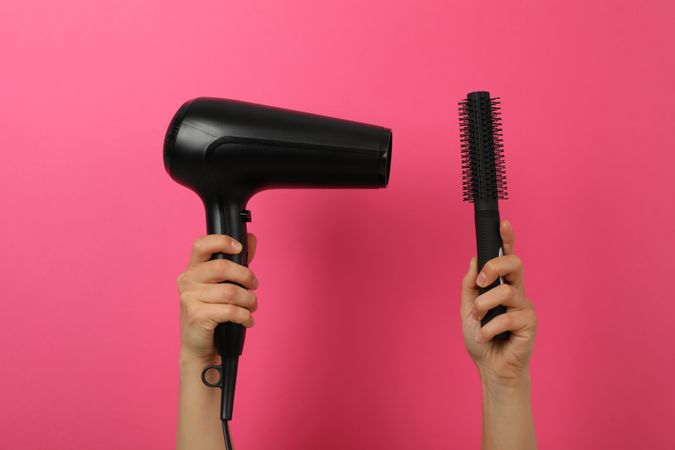 Female hands hold hair dryer and hair brush on pink background