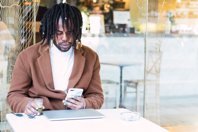 Black man with laptop browsing on mobile phone on restaurant terrace