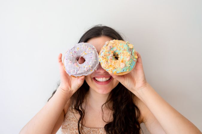 Playful woman making glasses of donuts while standing by white wall