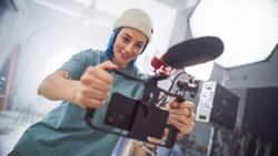Young Female Cinematographer With Blue Hair Adjusts Camera Equipment In Modern Studio, Focusing On Creative Video Production For Advertising, Showcasing Teamwork And Artistic Skill.