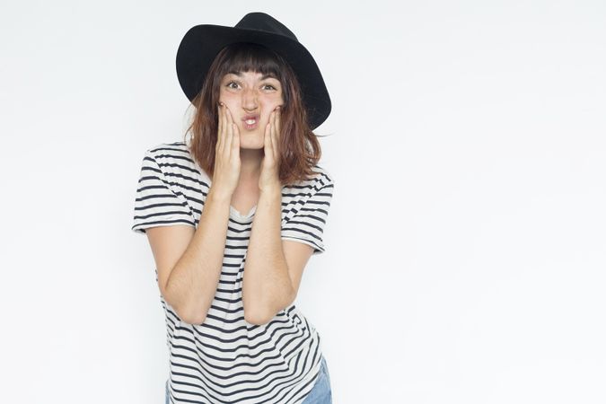 Female in striped shirt and felt hat holding mouth making funny face, copy space