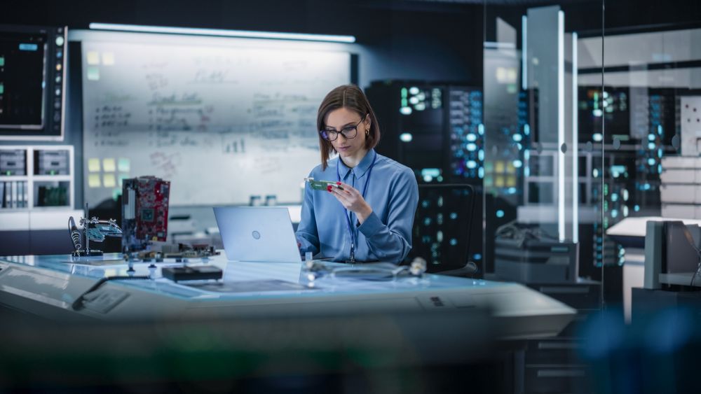 Portrait of Female Specialist Using Laptop Computer to Improve Electronic Circuit Board in Modern Office. Professional Engineer Thinking and Typing Ideas for Artificial Intelligence Project