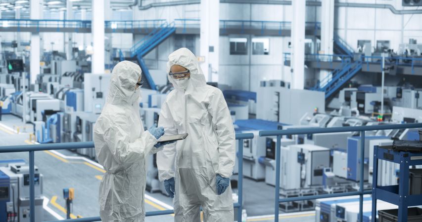 Two Female Technicians in Protective Coveralls Having a Conversation and Using Tablet Computer. Specialists Monitoring, Analyzing Conditions at a Modern Electronics Factory with Automated Robots