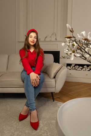 Teen girl in red blouse sits elegantly on sofa indoors.