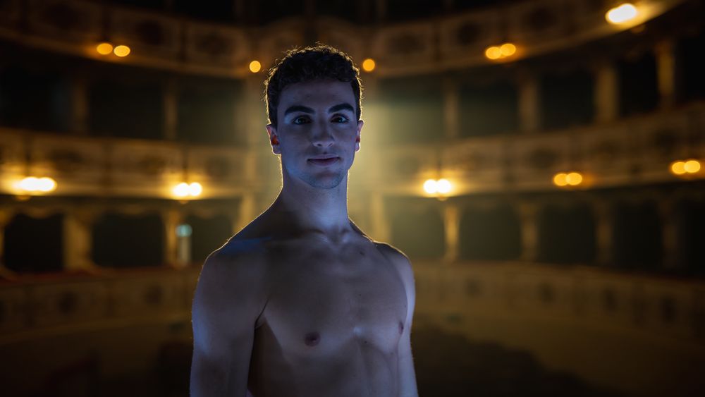 Cinematic Shot of Young Graceful Male Classical Ballet Dancer Looking at the Camera and Smiling in Classic Theatre with Dramatic Lighting.
