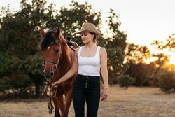 Young woman in hat walking with her horse in the countryside at sunset