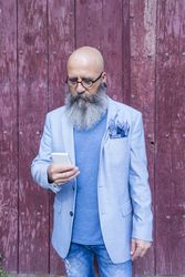 Front view of a mature trendy bearded hipster standing against a red vintage door outdoors while using a mobile phone
