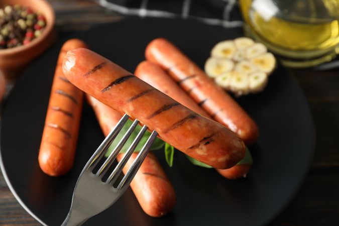Close up of grilled sausages or hot dogs on a fork on dark ceramic plate with garlic