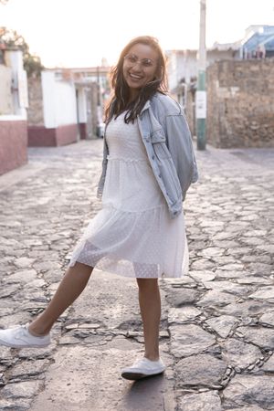 A young Hispanic woman with sunglasses is smiling posing in the street during the sunset