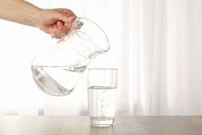 Hand pouring water in glass from pitcher in bright room