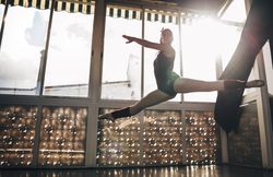 Young slim ballerina jumping above ground in studio flexing legs.