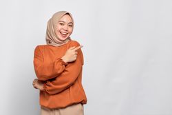 Smiling Muslim woman in headscarf and orange sweater pointing finger to blank copy space on her side