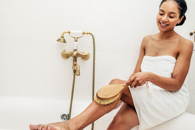 Woman wearing a towel sitting on edge of bath exfoliating her legs with a brush