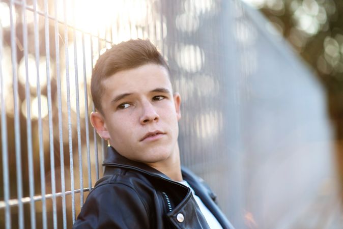 Teenage male wearing a leather jacket leaning on fence outside looking to his side