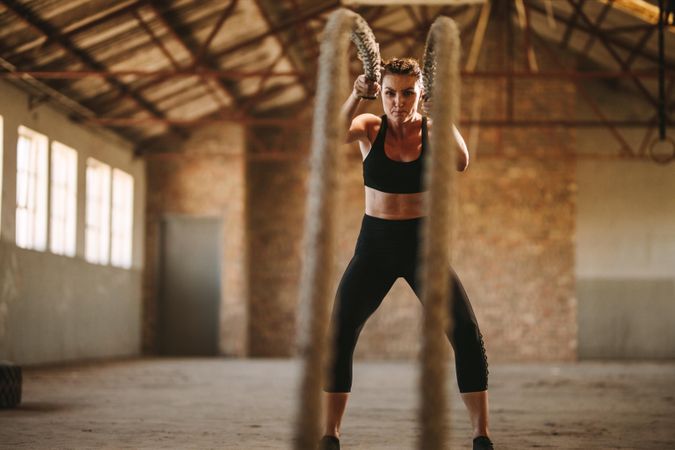 Strong woman working out with battle rope inside abandoned factory shade