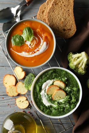 Two bowls of orange and green soup on wooden table with crackers and vegetables, vertical