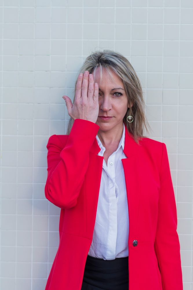 Portrait of a blonde woman wearing red jacket against a tiled wall covering her eye