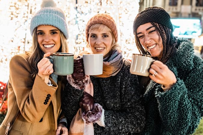 Three happy friends holding mugs and looking at the camera at a Christmas market.